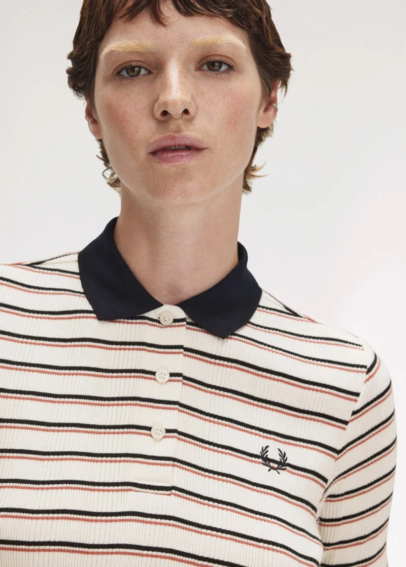Female model with short haircut and bleached brows wears ecru knitted polo shirt with black collar and think black and coral stripes