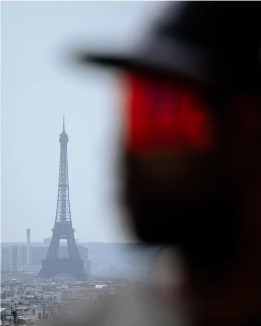 A photograph. In the foreground but out of focus is a headshot of a cyclist wearing a helmet with a red visor. The Eiffel Tower is in sharp focus in the distance.