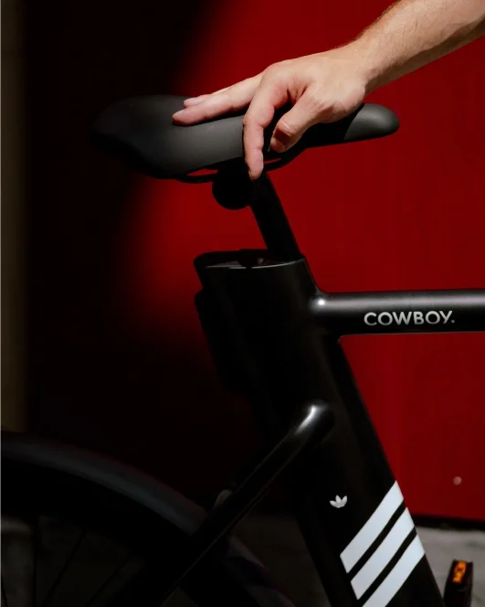 A photograph. A close up of a Cowboy bike, the dual Cowboy and Adidas branding is visible on the frame, a woman's hand rests on the saddle.