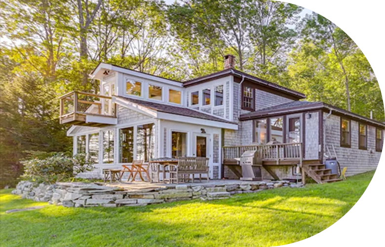 Two-story cottage with large windows, multiple decks, and a stone patio set beside a grassy yard.