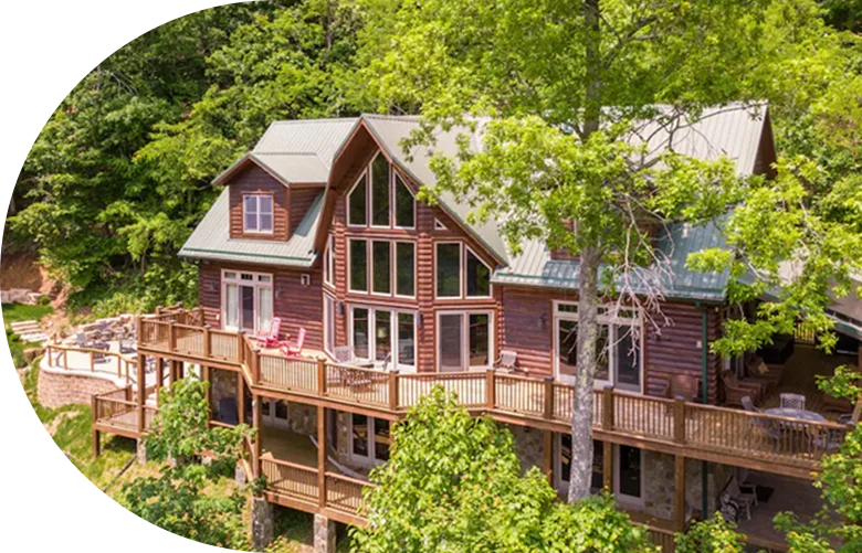 Large upscale cabin with wraparound wooden deck and tall windows set among dense forest.