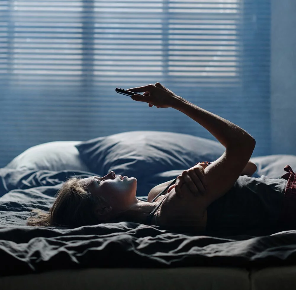 A young woman lying in bed with a serious expression while looking up at her phone