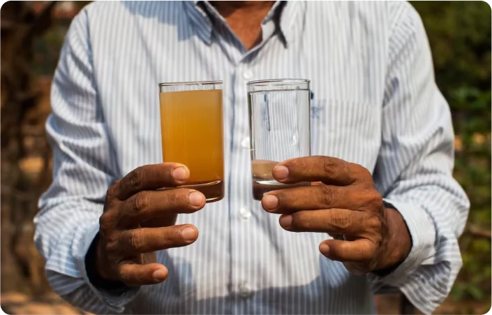 Photo of a person holding up a glass of dirty water from the previous water source and a glass of clean water from the current water source
