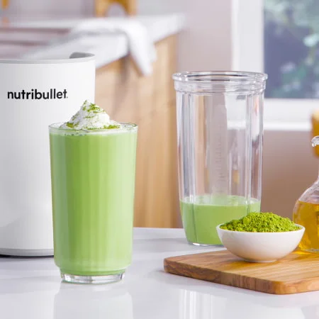 Green matcha smoothie in a nutribullet cup on a countertop, paired with fresh matcha powder and wooden utensils.