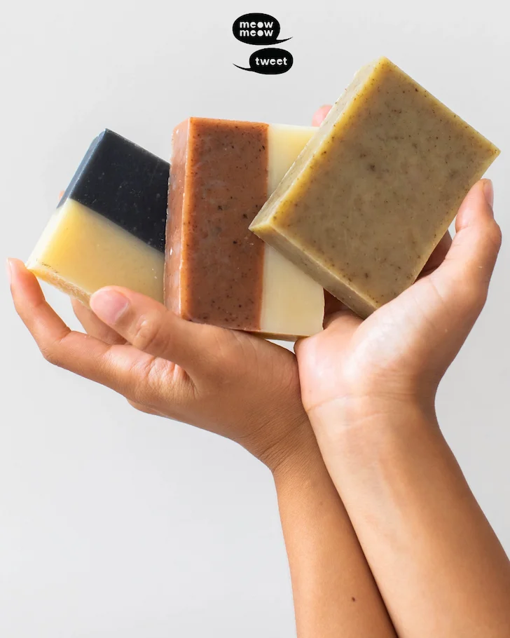 A pair of hands hold up three different bars of soap against a white backdrop.