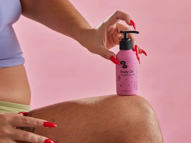 A person balances a pink bottle of body oil on their knee, they grasp the bottle with their long red nails.