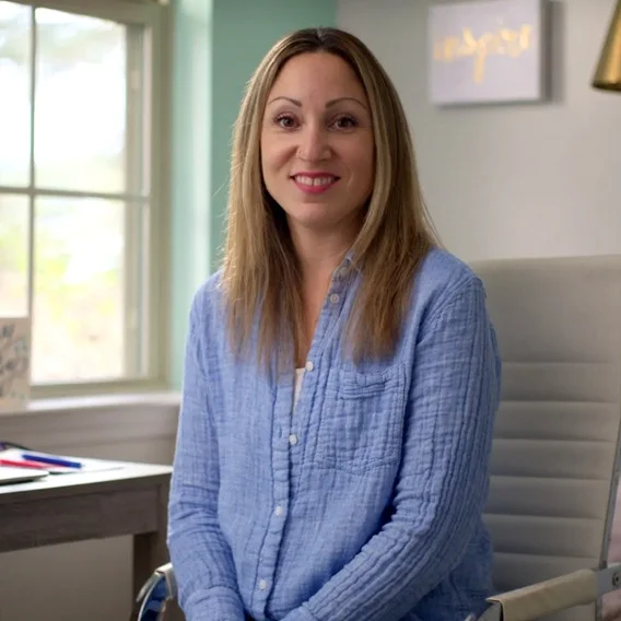 A woman with long, straight, light brown hair is seated in a chair and smiling. She is wearing a light blue, textured shirt. The background includes a window, a desk with papers and pens, and a picture frame with the word 'inspire.'