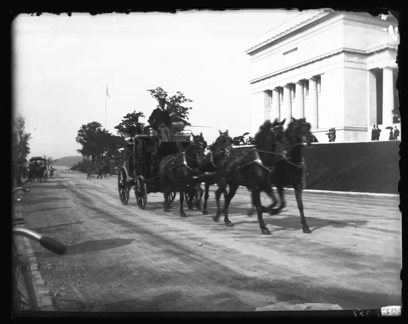 A black-and-white photo of a man driving a horse-drawn carriage past a large building