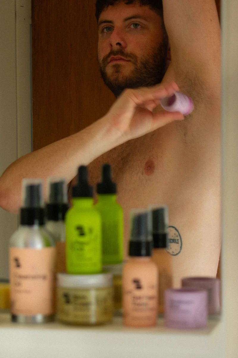 A white person with a beard applies a purple deodorant to their armpit