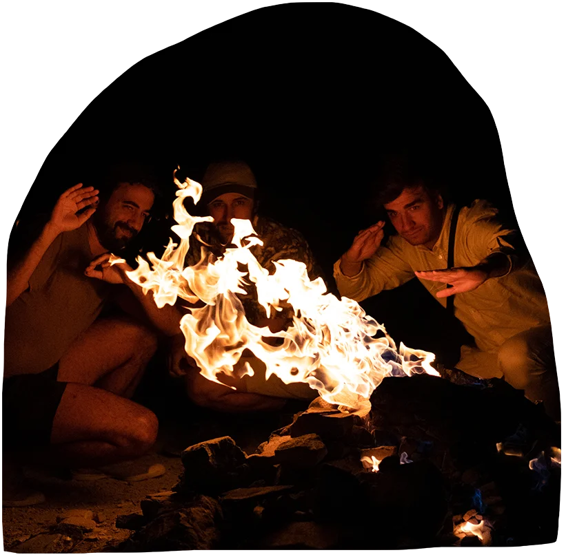 Three men hold their hands up against the flames of a small campfire while laughing