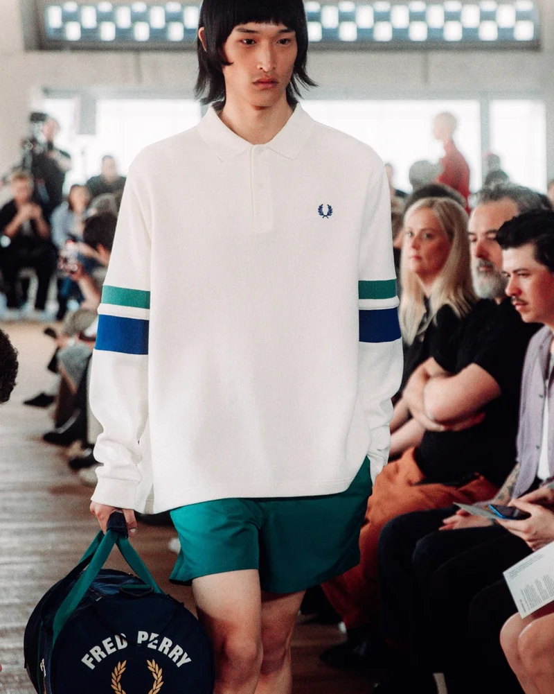 Male Asian model with fringe and jaw-length hair walks the runway in our long sleeve polo shirt with colour blocking stripes on the arms while holding a navy sports barrel bag