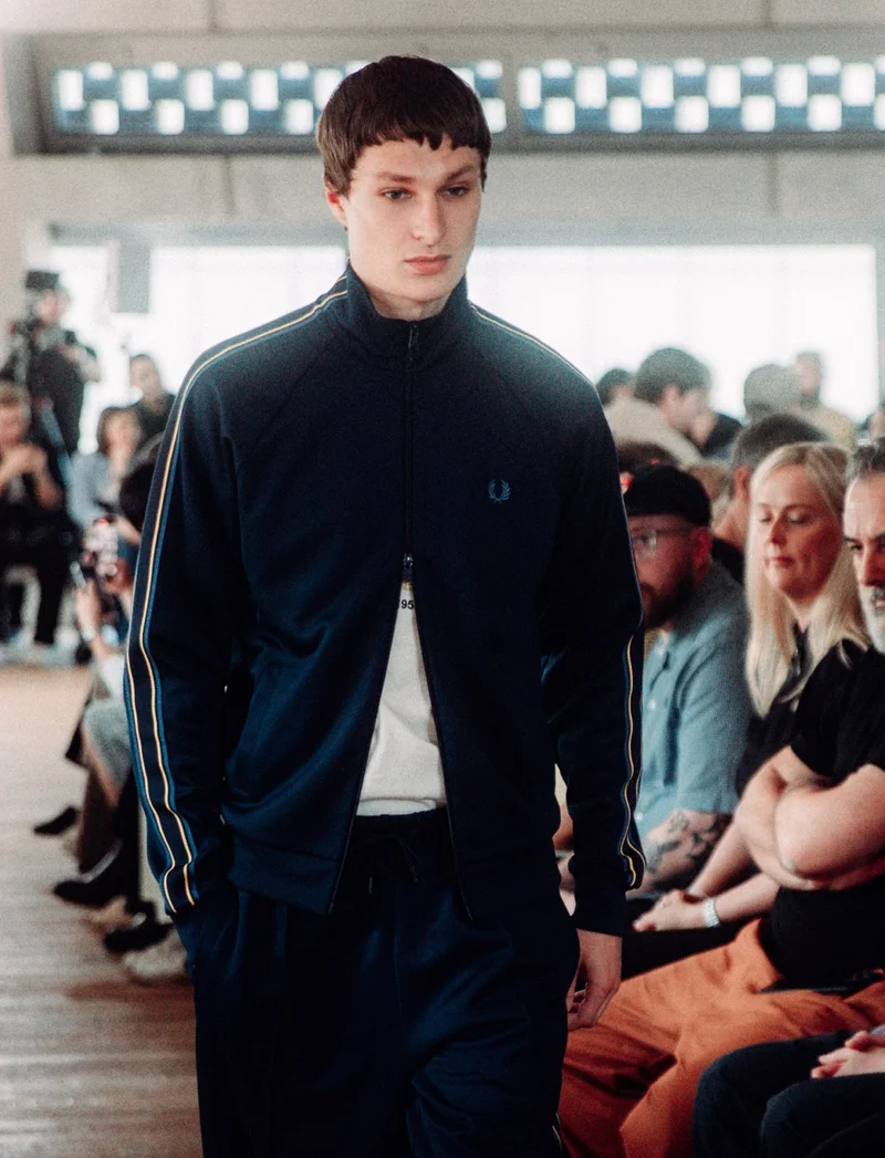 White male model with short brown hair walks the runway wearing our full navy tracksuit with gold, blue and black lined taping