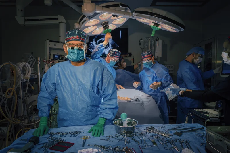 A doctor in blue scrubs looks at the camera. There are medical instruments on a table in front of him. In the background there are more doctors preparing for a surgery.