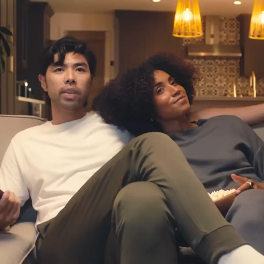 A man and a woman sit together on a couch; the woman rests her head on the man's shoulder.