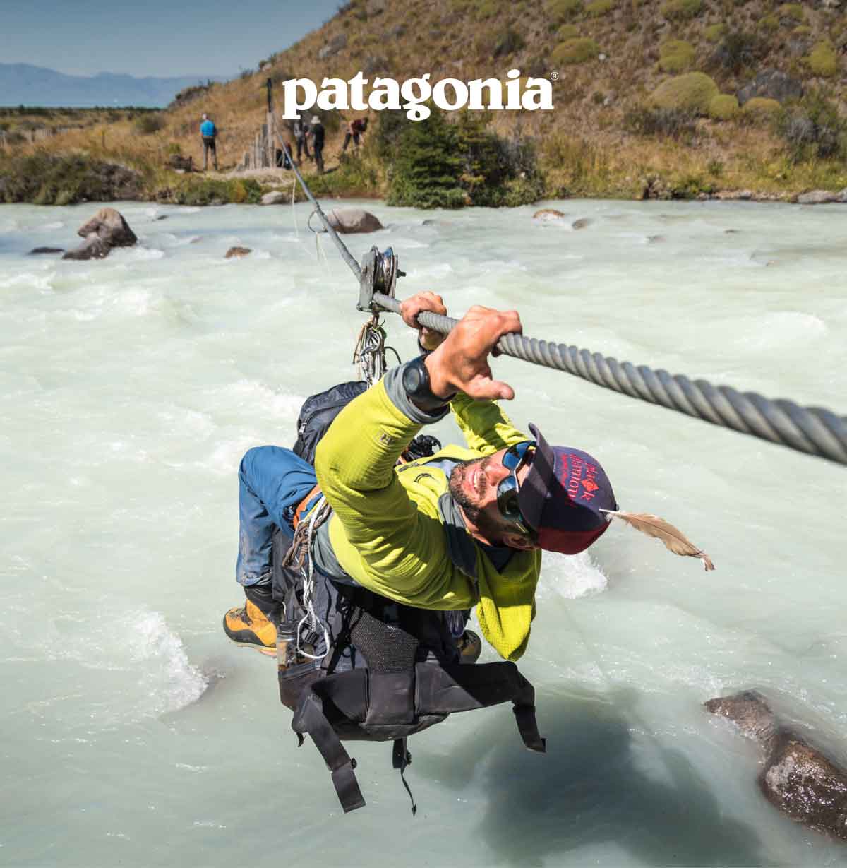 Patagonia. A mountaineer moves across a glacial river going hand over hand while dangling below a cable.