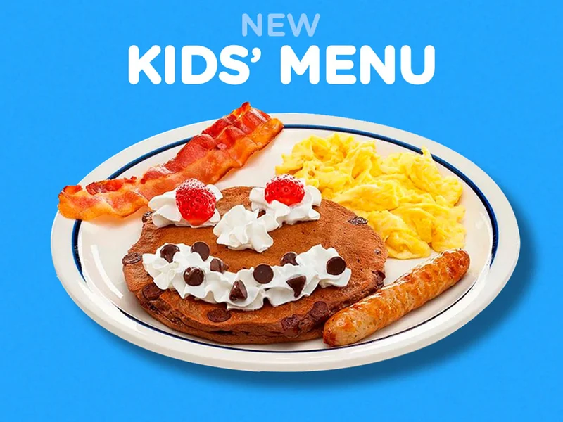 An image of the Happy Face Combo - a pancake with strawberry eyes, and a whipped cream nose and mouth, with scrambled eggs and a strip of bacon and sausage link.
