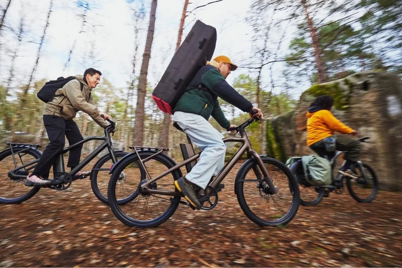 Three cyclists riding Cowboy e-bikes through a forest trail. The riders wear outdoor clothing, with one carrying a large padded backpack. The bikes feature integrated batteries and rear carrier racks, designed for off-road adventures. The background shows tall trees and moss-covered rocks.