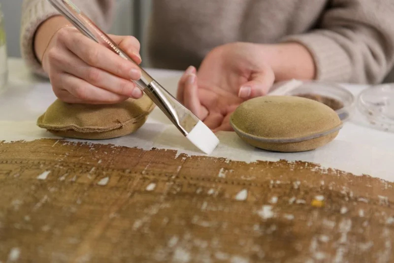 A close-up of two hands approaching papyrus paper with a brush