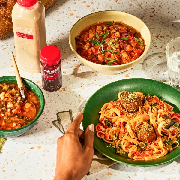 A variety of Splendid Spoon products arranged on a stone table. A hand reaches up from the bottom of the frame, about to pick up our Meatball Marinara Noodles