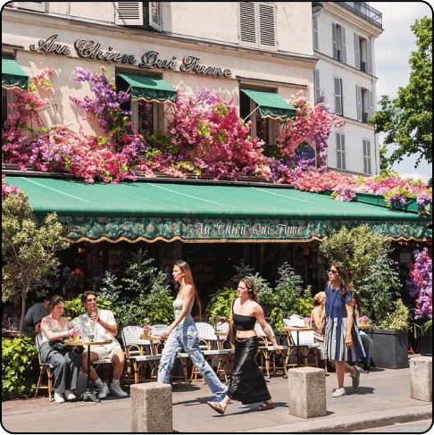 Image Description: People walk past a sidewalk cafe with a green awning and colorful flowers up the sides of the building.