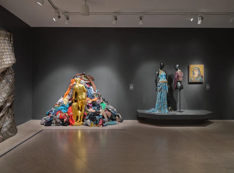 In a gallery of gray walls, a golden statue stands before a large pile of colorful clothing