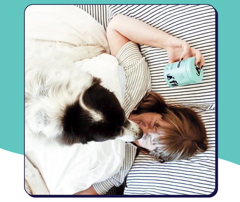 Dog jumping on human in bed with Finn Calming Aid tin in hand