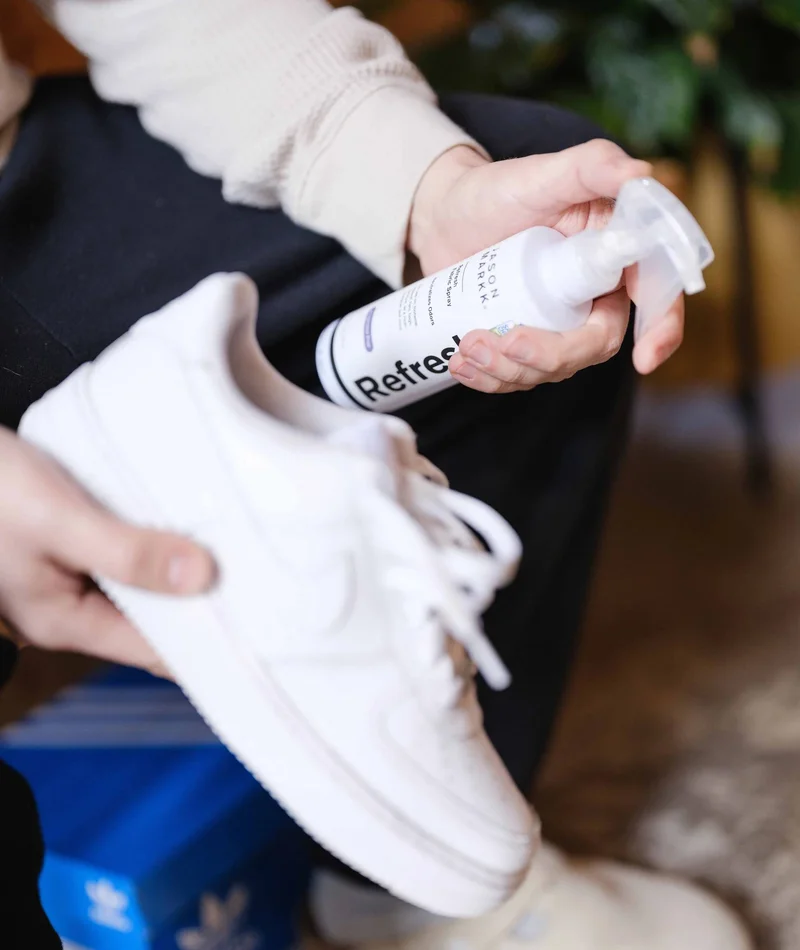 A medium shot shows a person holding a white sneaker and a white spray bottle labeled 'Refresh' with text including 'JASON MARK' and 'Fabric Spray' an...