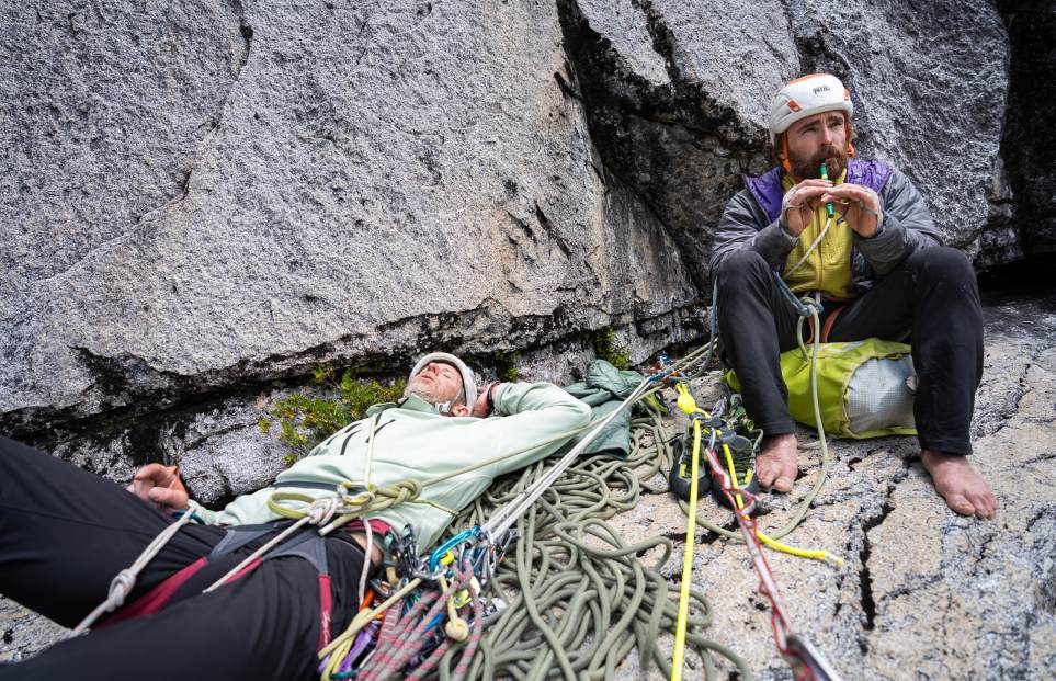 Two climbers on a belay ledge, one laying down and one playing a tin whistle.