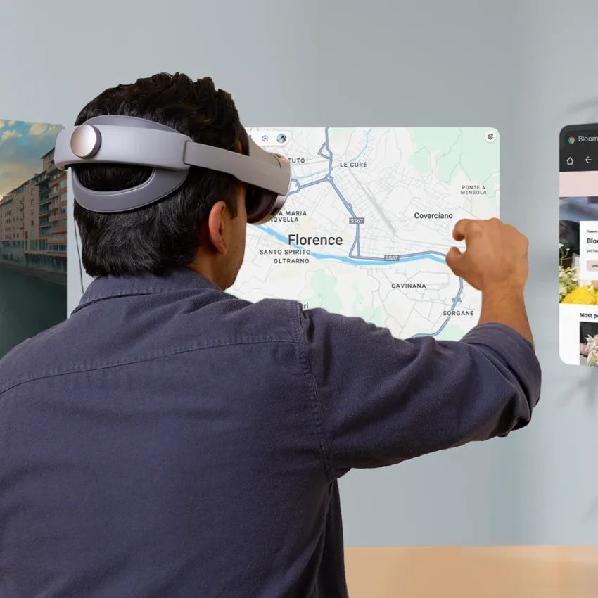A person from behind, wearing a grey XR headset, stands at a desk and interacts with several floating digital screens. The central screen is a large map of Florence,Italy, which they are pointing at with their right hand. Other screens to the left and right show photos.