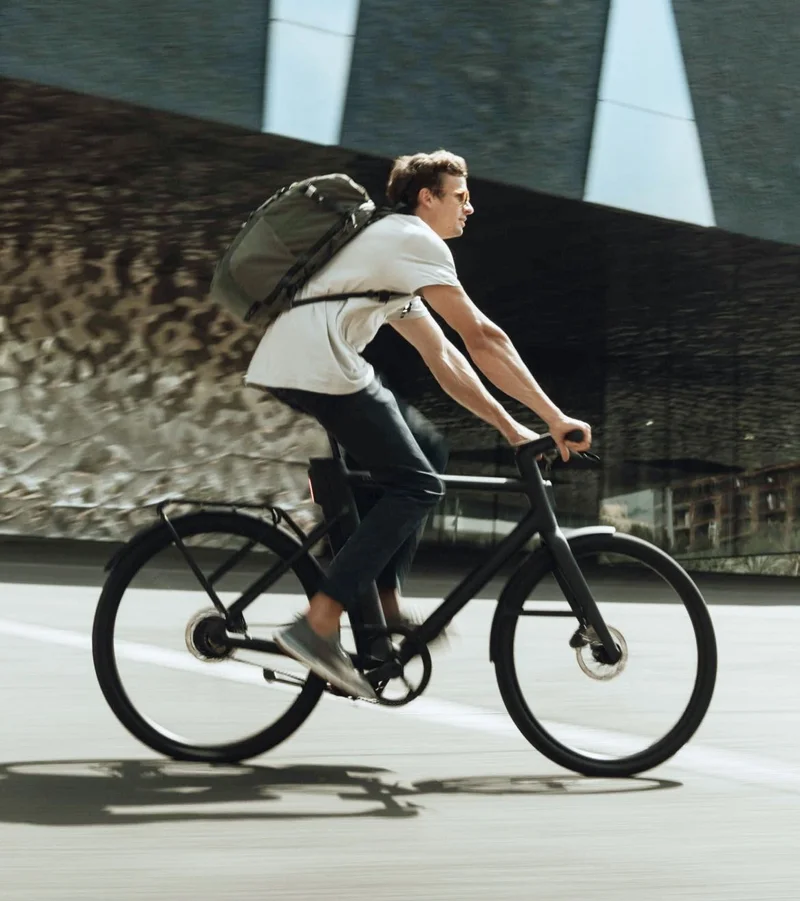 A photograph. A man wearing a rucksack rides a black Cowboy Classic in an urban setting