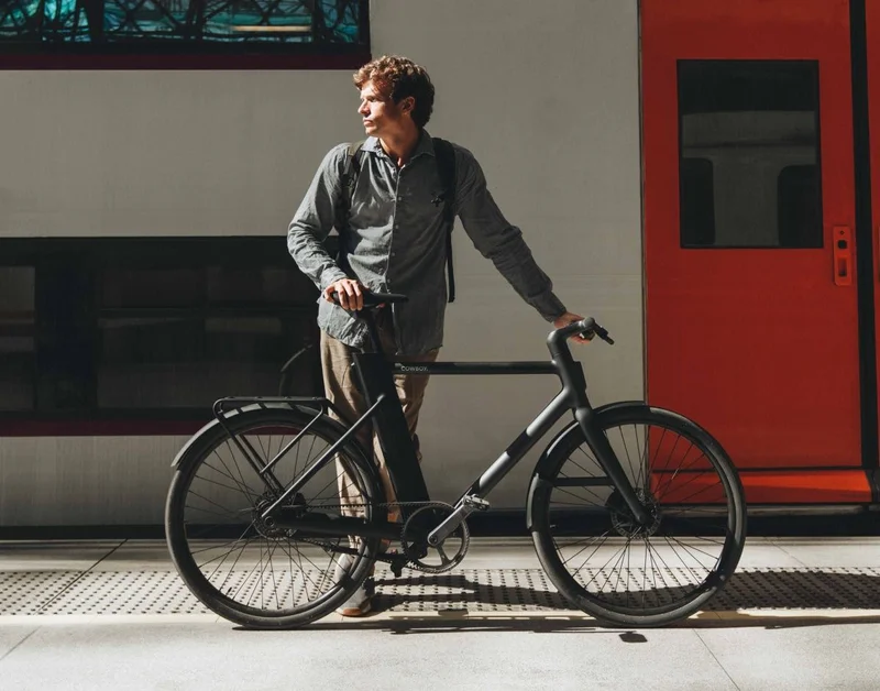 A photograph. A man stands with a black Cowboy Classic, a commuter train stands at a station platform behind him.
