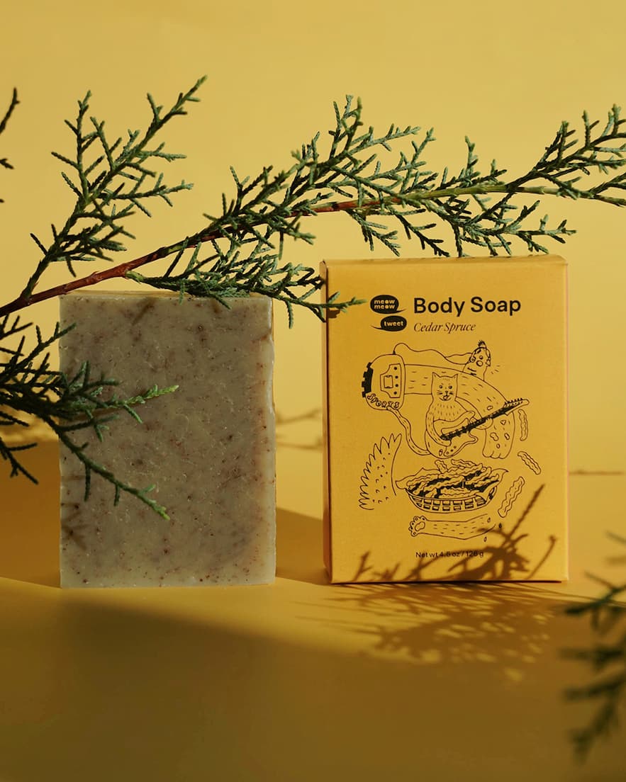 Two bars of soap sit on a yellow background with a juniper sprig in the foreground