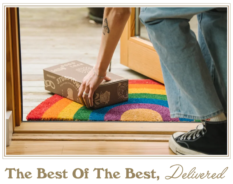 A person picks up a delivered Stumptown Roaster’s Pick subscription coffee box from a rainbow doormat at a front doorstep, with the headline “The Best Of The Best, Delivered.”