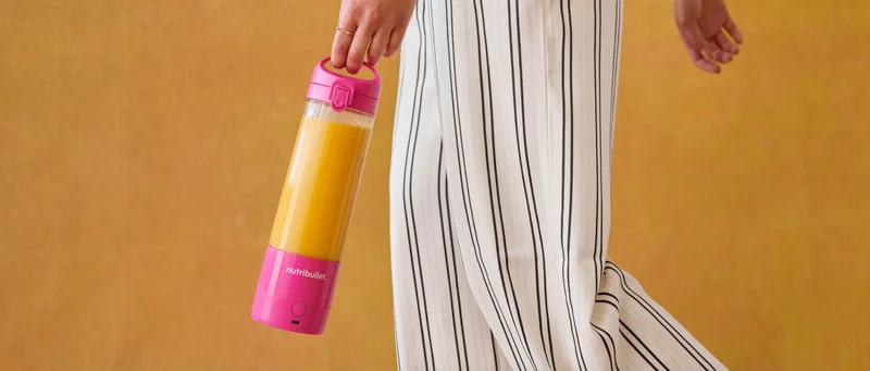 A close-up image of a person in striped pants holding the pink nutribullet Portable Blender containing orange juice against a warm background.