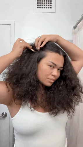 A person with long brown curly hair separates their hair and applies oil to their scalp