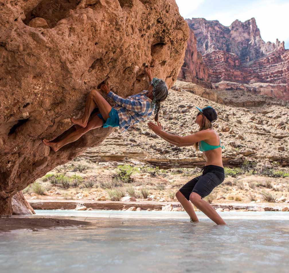 A person climbs a boulder over shallow water.