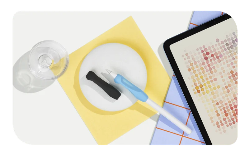 Paperlike's Pencil Grips in Charcoal and Paperlike Blue surrounded by paper, a glass, and an iPad.