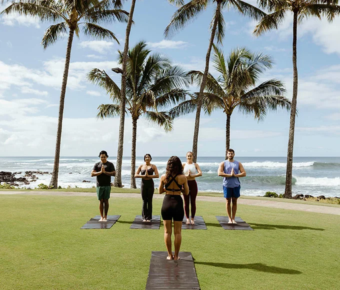 A female yoga instructor is standing in front of four people standing on yoga mats, on the grass. A beach and palm trees are in the background.