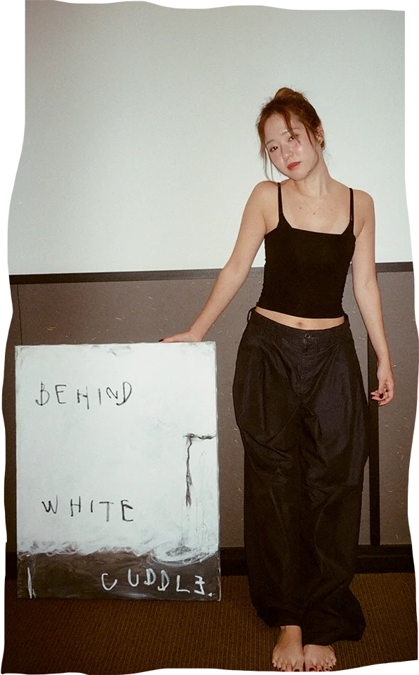 Artist Momo standing barefoot in a relaxed pose, wearing a black crop top and loose black pants, next to a canvas with the words 'BEHIND WHITE CUDDL3' painted on it.