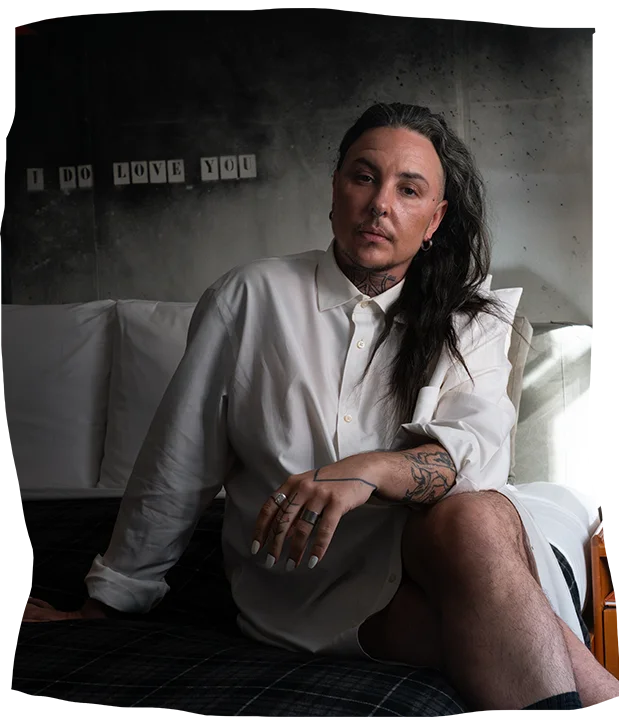 Performance artist S.J Norman, with long dark hair, sitting contemplatively on a bed in a dimly lit room. They wear a crisp white shirt, display visible tattoos on their hand, and are set against a raw concrete wall with framed pictures.