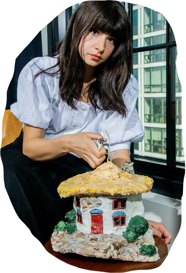 Artist Janie Korn seated by a window, holding one of her intricately carved wax candles resembling a house with red doors and a yellow roof, surrounded by miniature trees.
