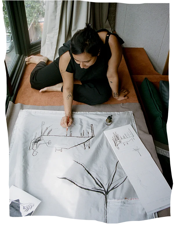 Artist Mia Boe seated at a window, intently working on a large sketch with drawings of abstract structures, while surrounded by scattered art supplies and paper.