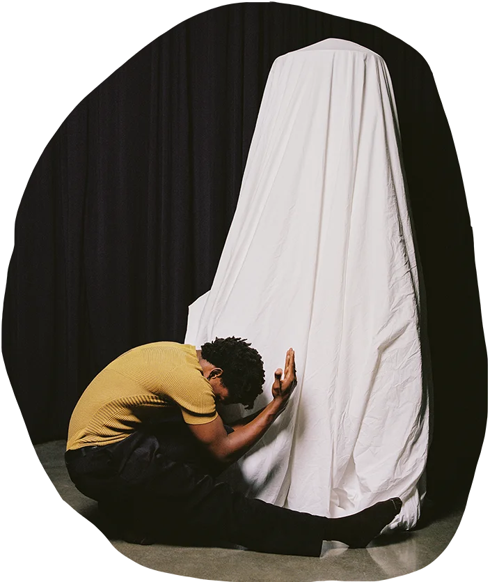 Artist Qwenga in a mustard-colored shirt, kneeling and embracing a draped white cloth that cascades from ceiling to floor, set against a dark background, conveying a sense of intimacy and connection.