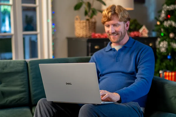 A parent siting on a couch in their living room doing their holiday shopping on a Dell laptop computer.