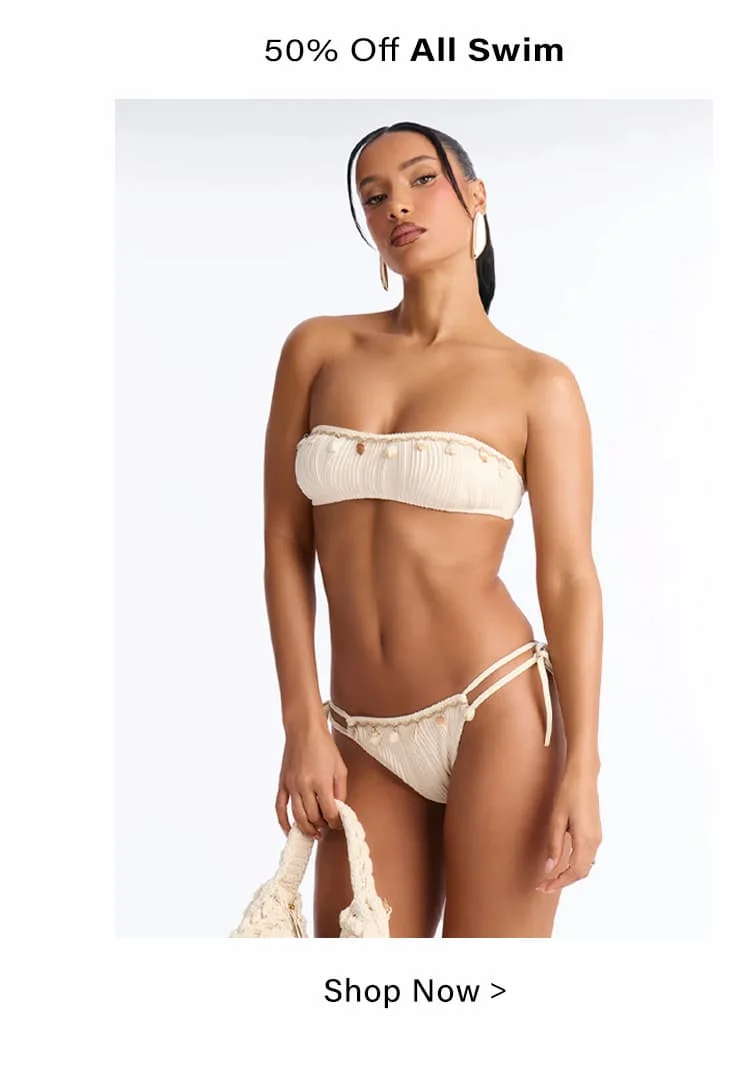 Cream textured bandeau bikini with tie-side bottoms and delicate embellishments, modeled by a woman holding a woven beach bag.