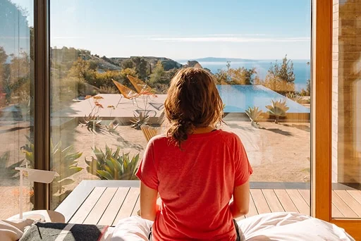 A woman sits on the edge of a bed, looking out over a sandy outdoor space with lounge chairs and a swimming pool.