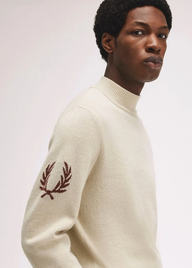 Man wears wool-blend long-sleeved high-necked knitted jumper in colour Ecru, with a large rust coloured laurel wreath knitted into the upper arms.