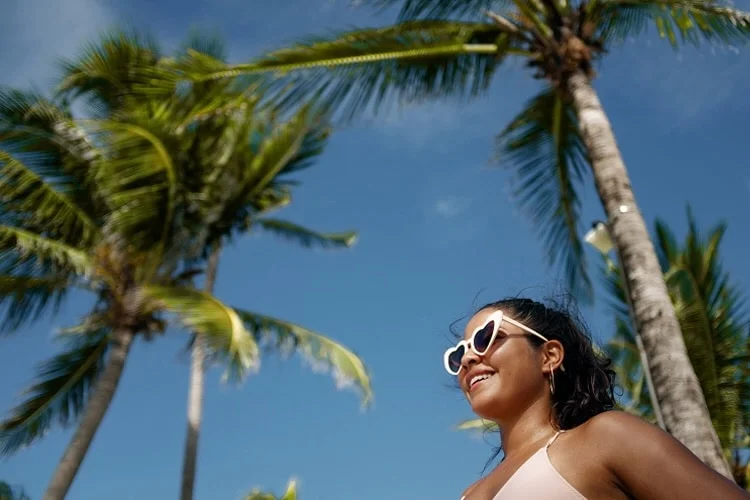 A woman in a pink bathing suit and sunglasses smiles with palm trees in the background.