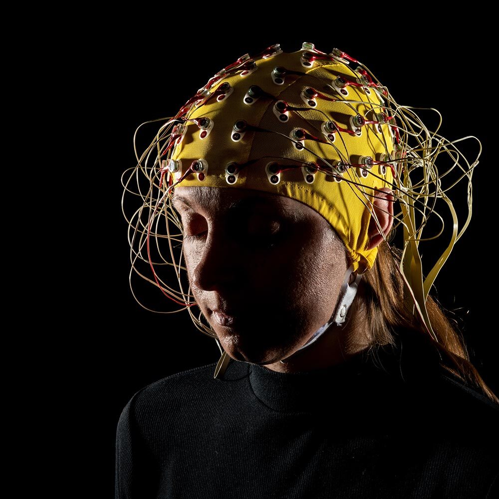 A woman wearing an electroencephalogram around her head sits in a dimly lit space with her eyes closed