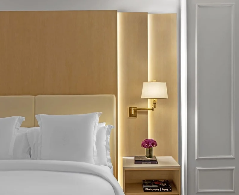 Image description: Hotel room with light wood walls and furnishings and white bed sheets. A small bedside table features a book on photography and a small vase with purple flowers.